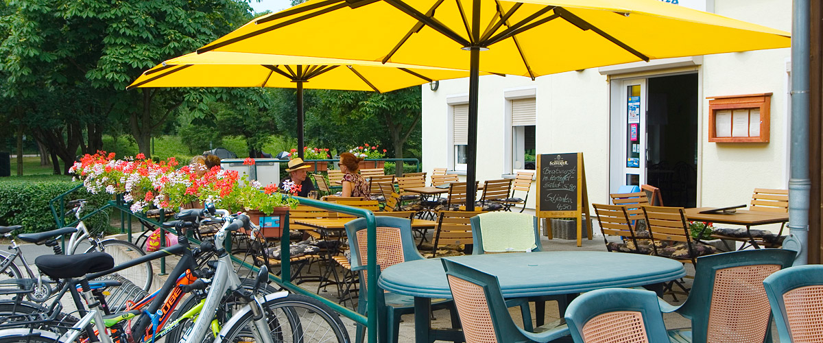Kleiner Außenbereich der Gaststätte mit Tischen und Stühlen, zwei gelben Sonnenschirmen sowie einem Fahrradabstellplatz.