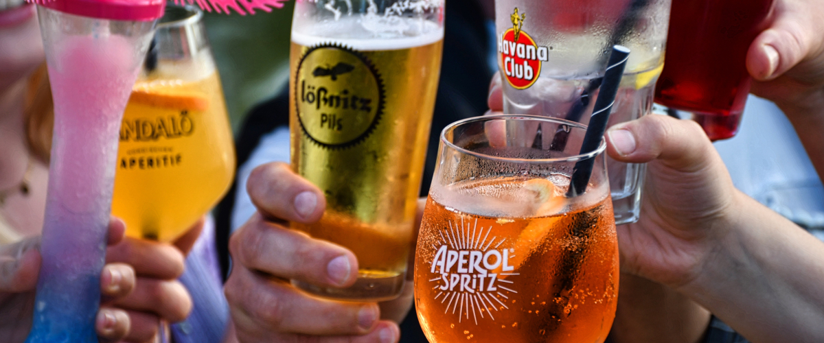 Mehrere Personen stoßen mit verschiedenen Getränken an, darunter Bier, Aperol Spritz, ein roter Longdrink und bunte Cocktails. Die Gläser werden in den Händen gehalten und vermitteln eine gesellige, sommerliche Atmosphäre.