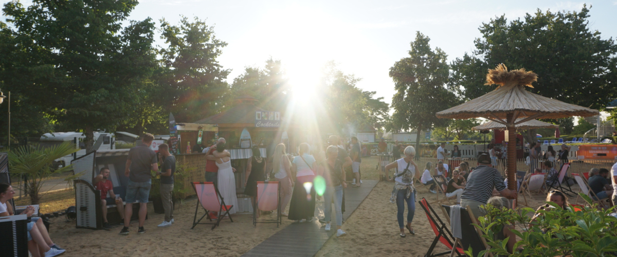 Strandbereich bei Sonnenuntergang mit Menschen, Sitzgelegenheiten im Sand, Sonnenschirmen aus Stroh und einem zentralen Holzweg. Im Hintergrund befindet sich eine kleine Cocktailbar, während die warme Abendsonne eine entspannte, sommerliche Atmosphäre schafft.