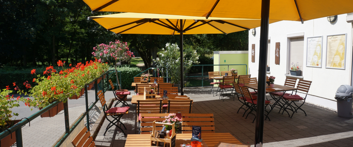 Außenterrasse eines Restaurants mit mehreren Holztischen und Stühlen unter großen gelben Sonnenschirmen. Entlang des Geländers stehen Blumenkästen mit roten Blumen. Die Terrasse liegt ebenerdig direkt am Gebäude und wirkt hell, freundlich und gut zugänglich.